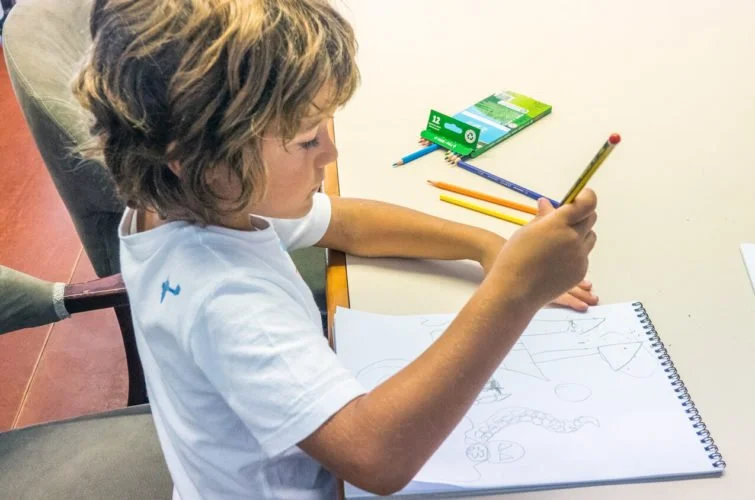 Niño dibujando en un cuaderno con lápices de colores y útiles escolares en la mesa.