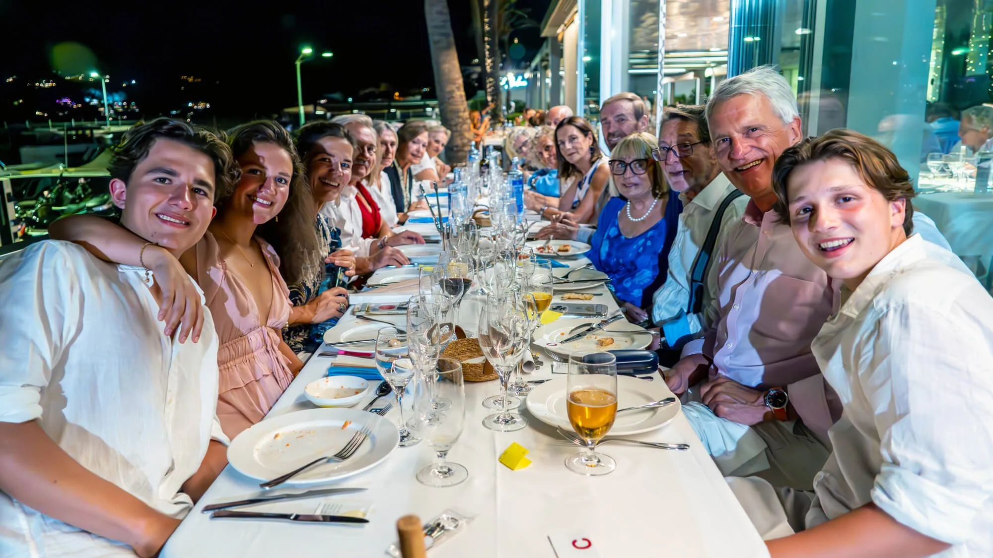 Grupo de personas en una cena al aire libre, sonriendo y disfrutando en una mesa con platos, copas y comida, en la noche