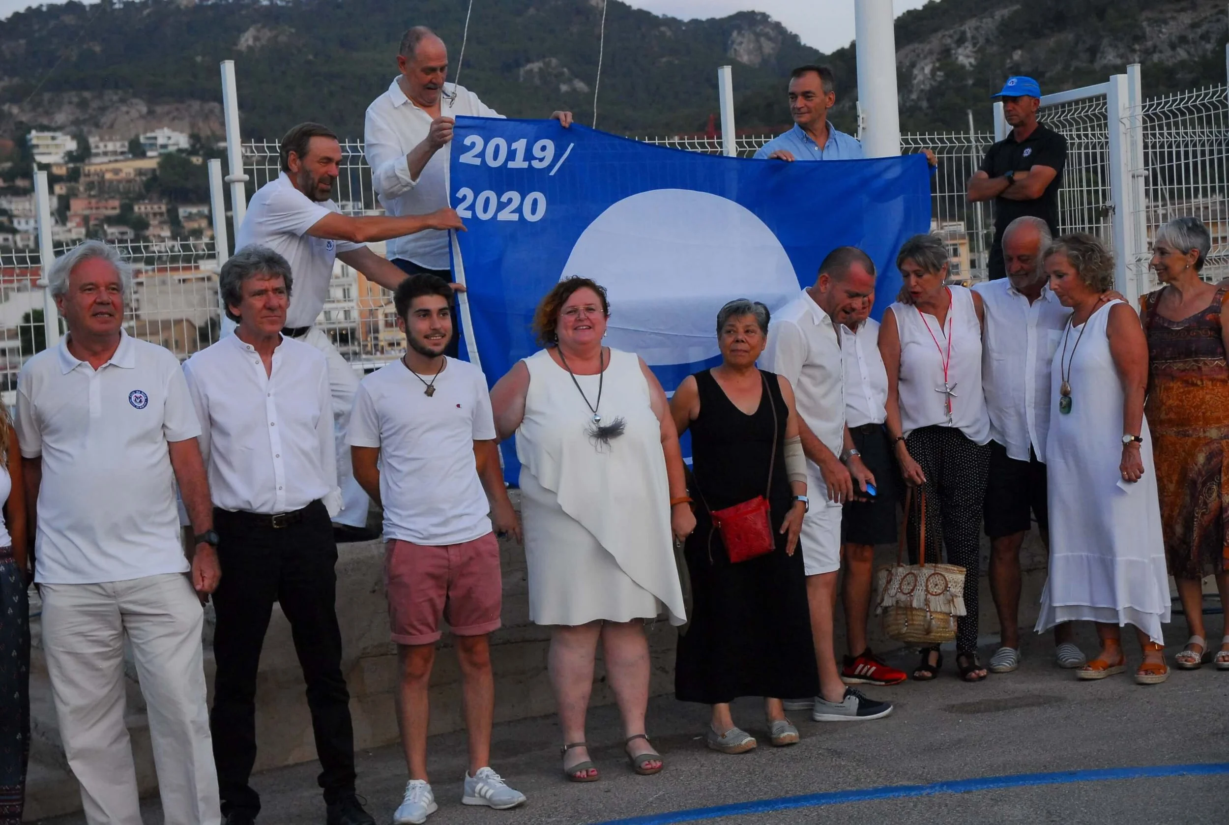 Grupo de personas en una reunión o evento, algunas sosteniendo una bandera azul con los años 2019 y 2020, en un lugar al aire libre con colinas o montañas de fondo.