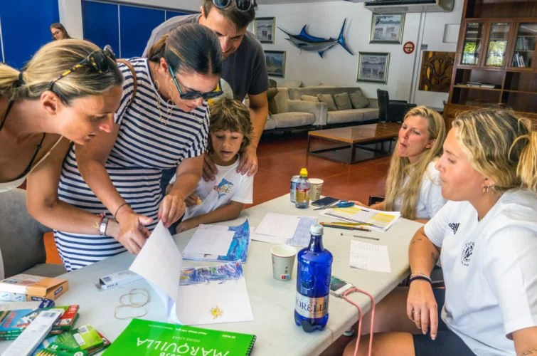Grupo de personas, incluyendo adultos y niños, revisando papeles y objetos en una mesa en un ambiente interior, con accesorios y libros distribuidos.