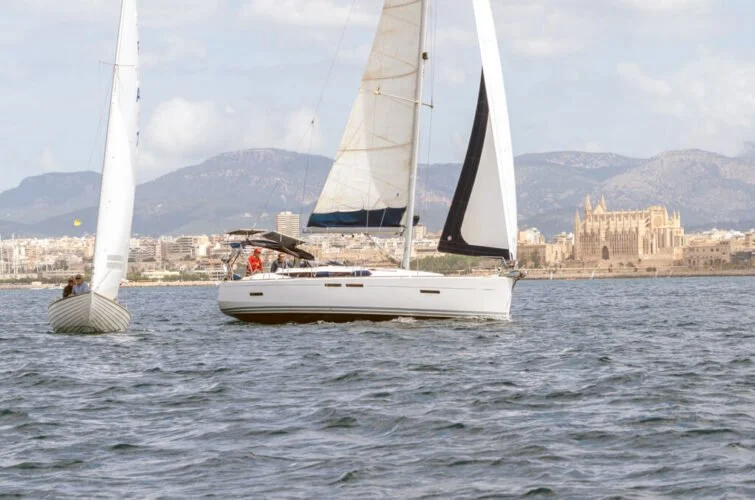 Barco de vela navegando en el mar con ciudad y montañas al fondo.