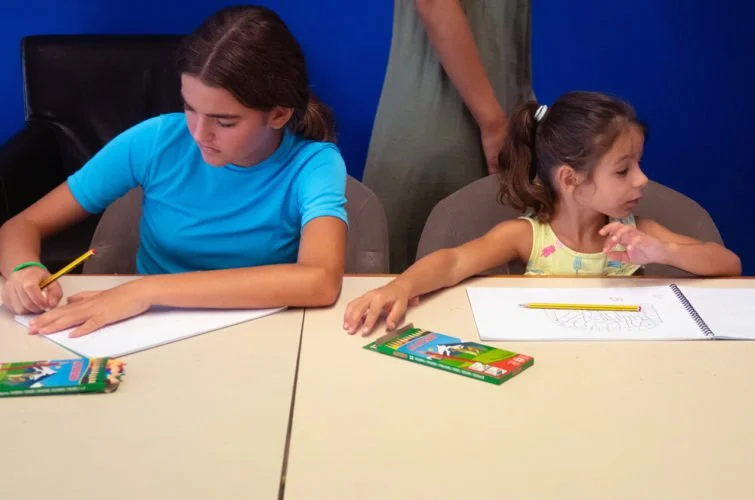 Dos niñas sentadas en una mesa, una más grande y otra más pequeña, haciendo tareas con lápices y cuadernos, en un entorno de aula o sala de clases.
