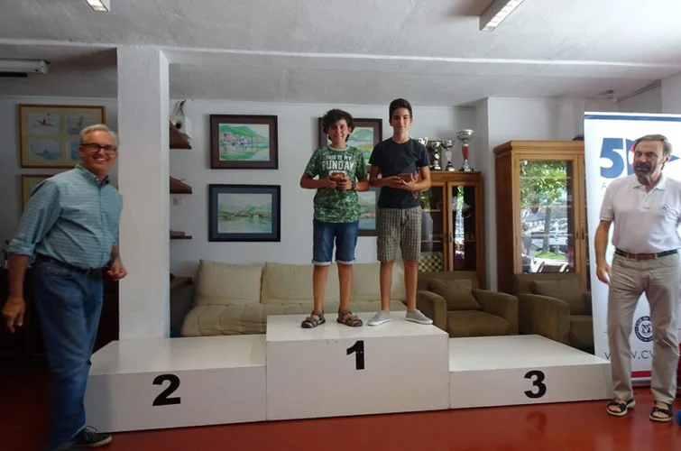 Dos niños de pie en un podio de premiación, en primer lugar, con un adulto a la izquierda y otro a la derecha, en un ambiente interior con cuadros en la pared y una vitrina con trofeos.