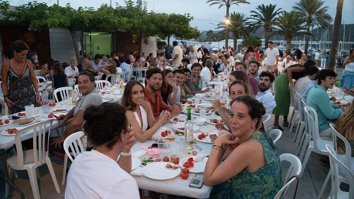 Grupo de personas en una comida o fiesta al aire libre, sentadas en una mesa larga con comida y bebidas, rodeadas de palmeras y un ambiente festivo.