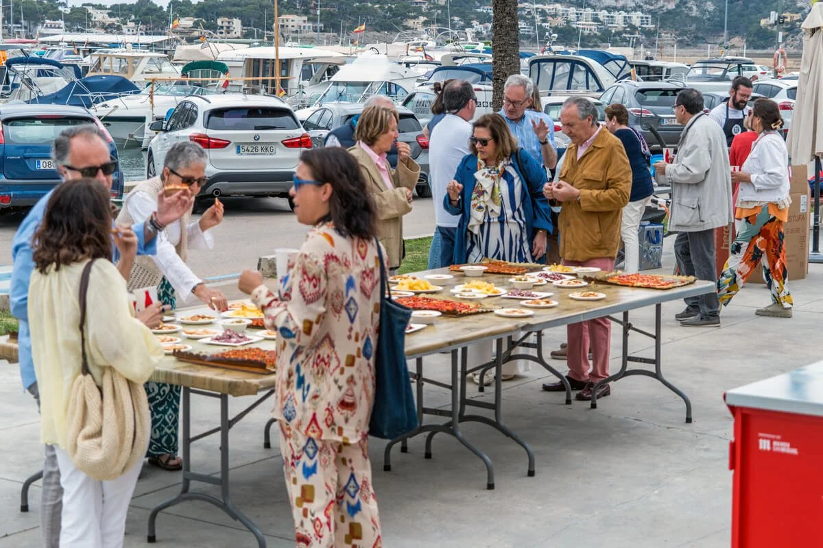 Un grupo de personas en un evento al aire libre, con mesas llenas de comida y una marina con barcos al fondo.