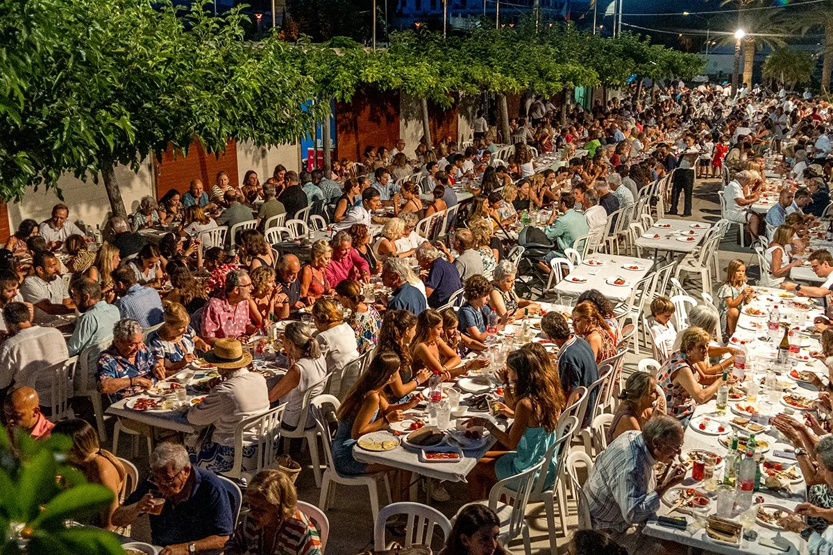 Una gran celebración al aire libre con muchas mesas largas llenas de personas comiendo, en un entorno nocturno.