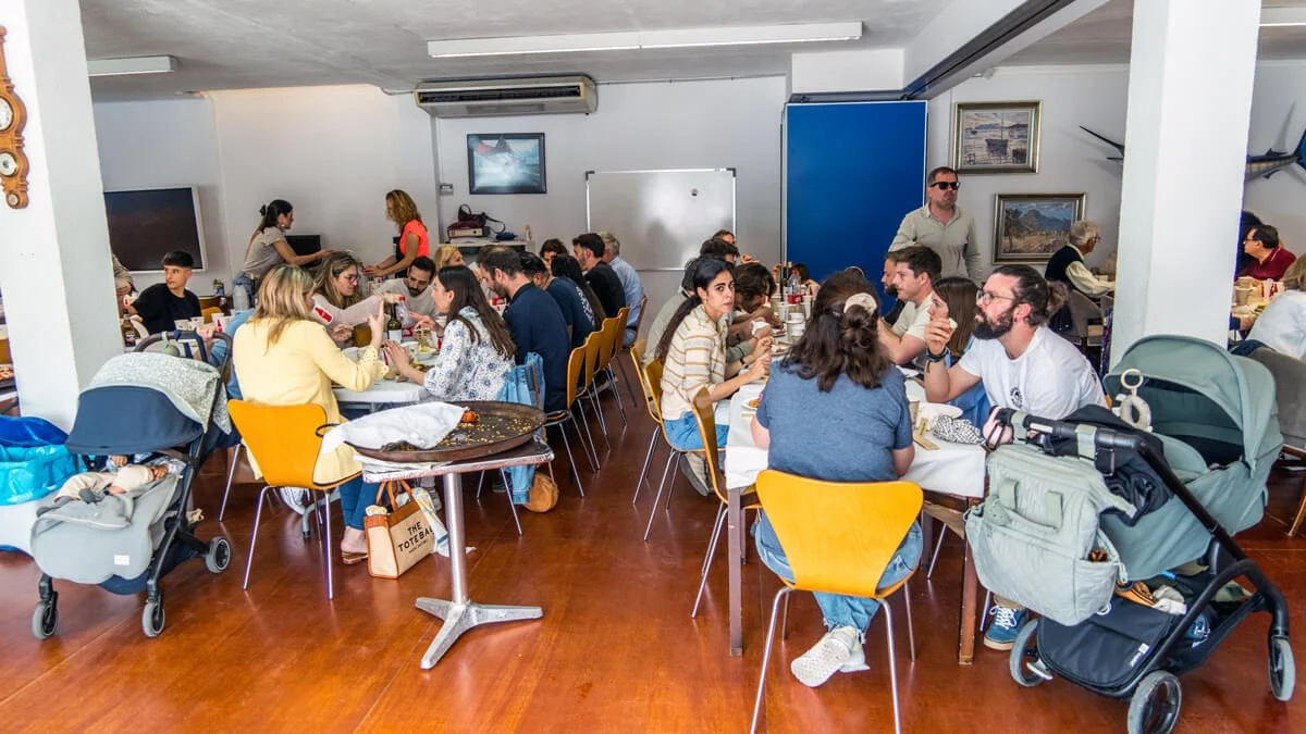 Personas en una comida grupal en un restaurante con paredes blancas, cuadros y una pizarra blanca, comiendo y conversando, con cochecitos de bebé estacionados.
