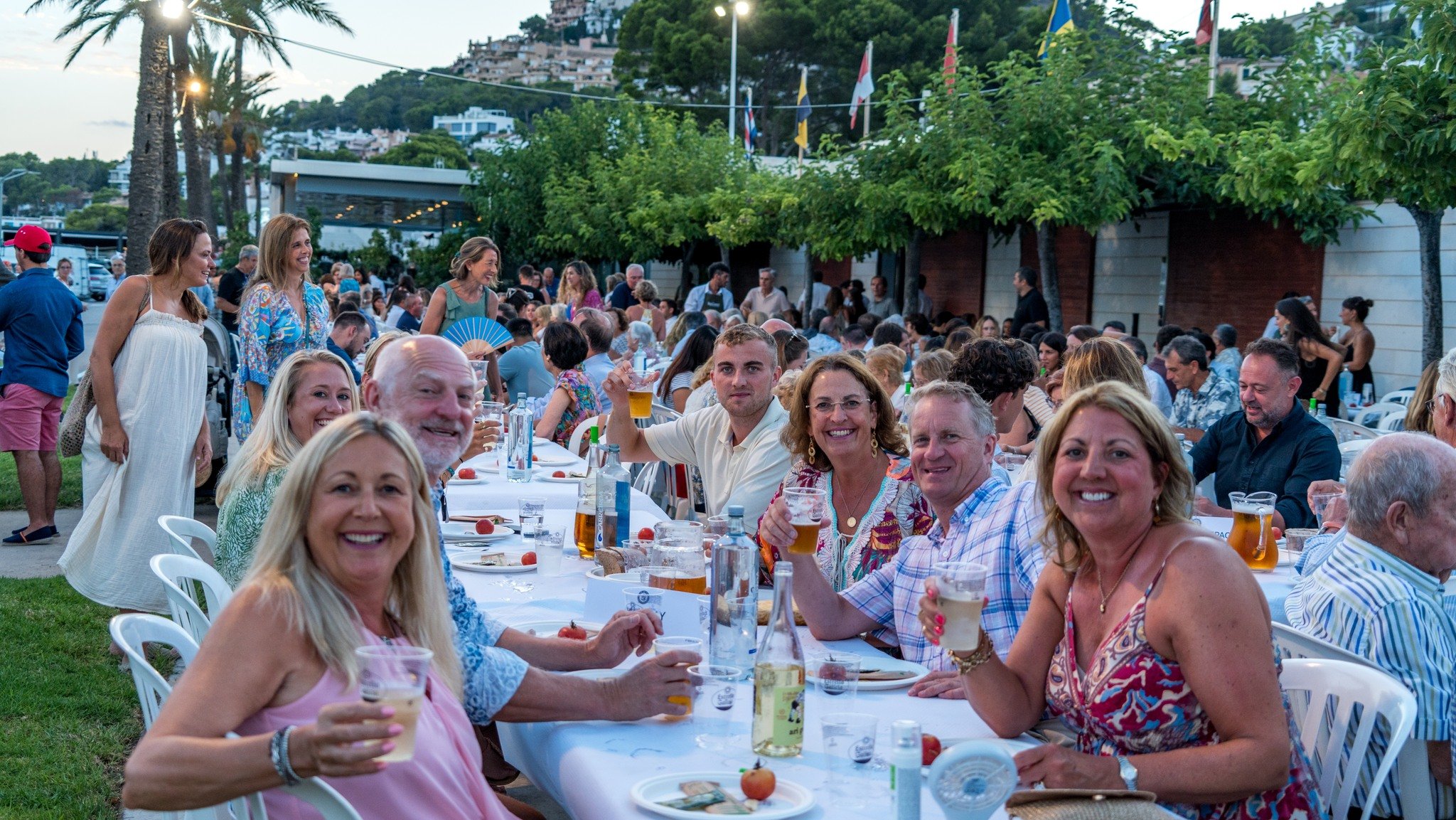 Un grupo de personas disfrutando de una comida en una fiesta al aire libre, con muchas sonrisas, bebidas y comida en las mesas, rodeados de árboles y banderas, durante el atardecer