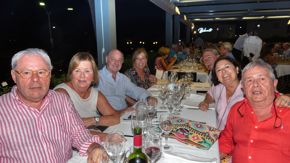 Grupo de personas sonriendo y disfrutando en una cena en un restaurante, con mesas y copas de vino.