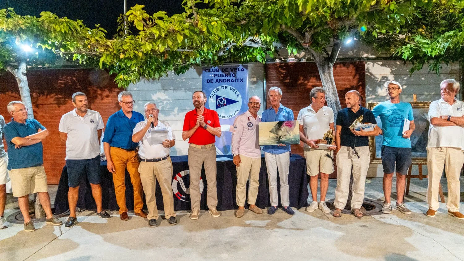 Grupo de personas en un evento al aire libre en la noche, con un árbol y un cartel que dice 'Club de Vela Puerto de Andraitx' en el fondo, algunas personas sosteniendo premios, una persona leyendo un discurso con micrófono, todos vestidas de manera c