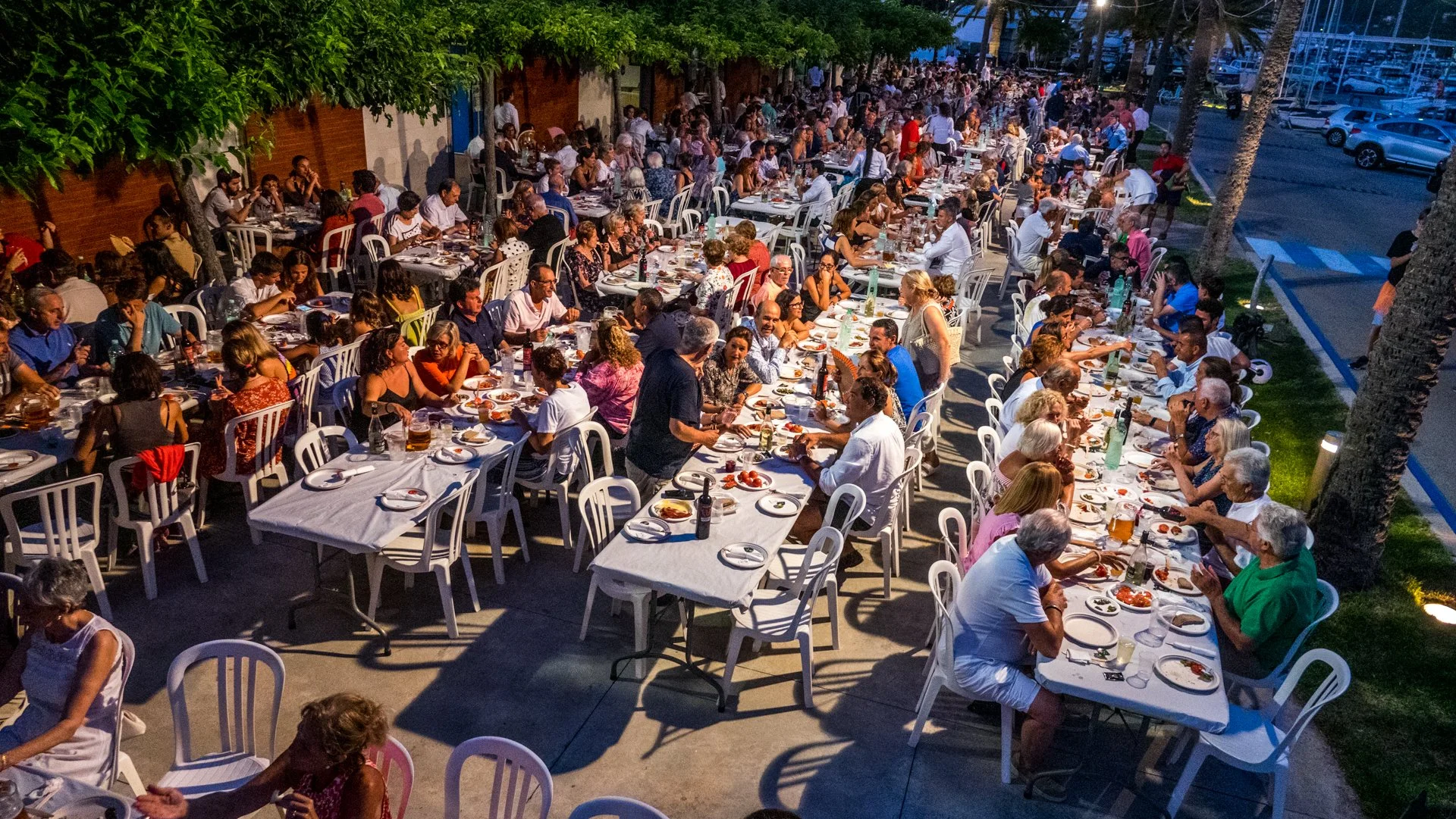 Escena de una cena al aire libre con muchas personas sentadas alrededor de varias mesas largas en la noche, disfrutando de comida y bebidas en un evento social.