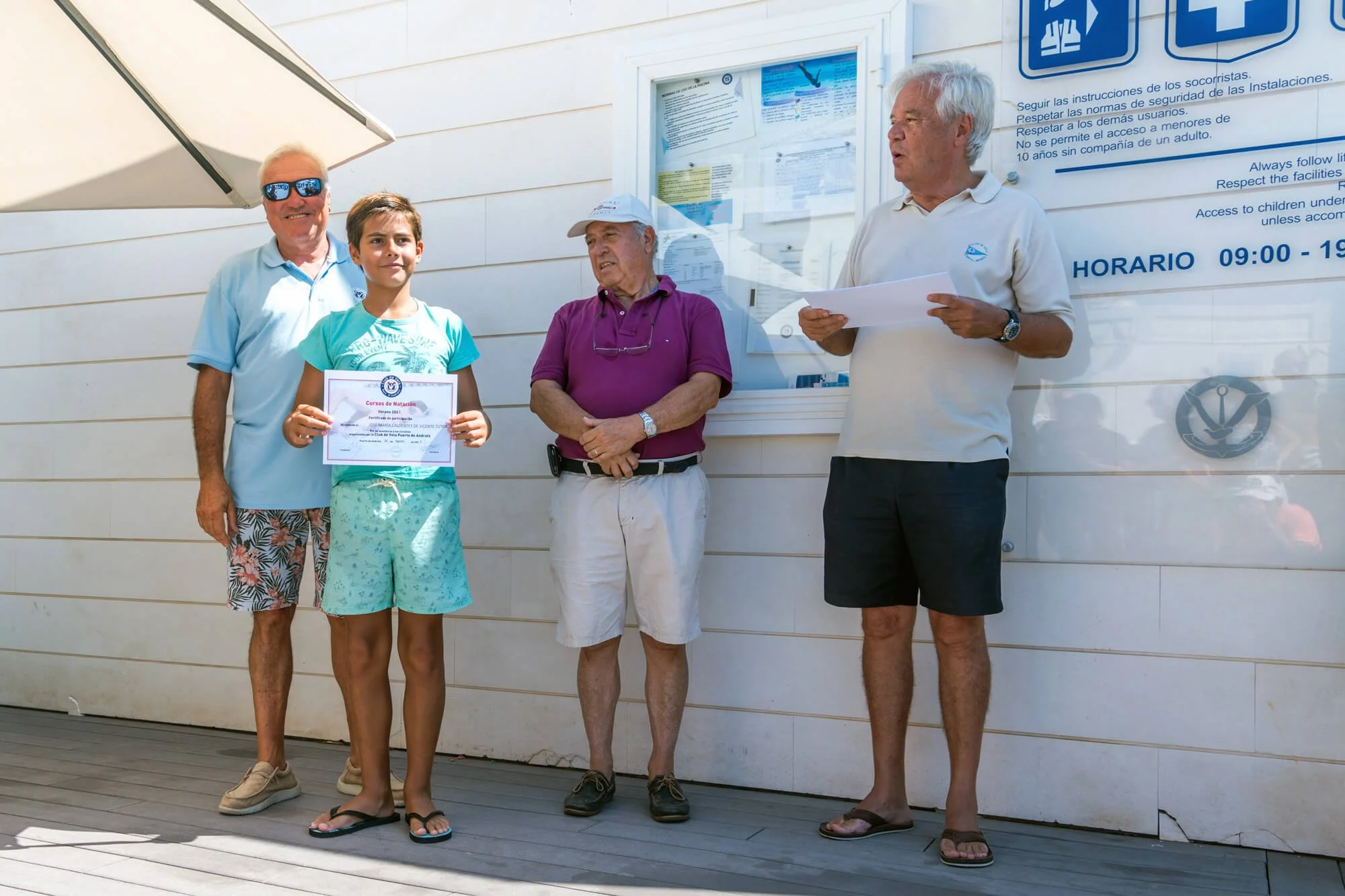 Cuatro personas en una terraza de un lugar de natación, donde una niña recibe un certificado y tres adultos la acompañan, mientras uno lee un papel. La escena es en un día soleado, y las personas están vestidas con ropa informal y Cómoda.