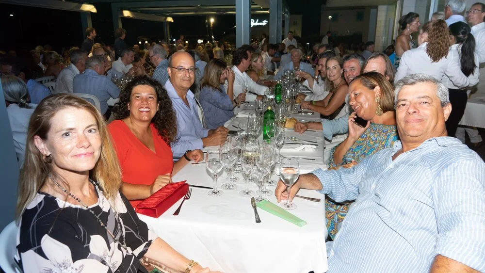 Grupo de personas en una cena elegante en un restaurante o evento social, sentados en una mesa larga con copas y platos, algunos conversando y otros sonriendo.