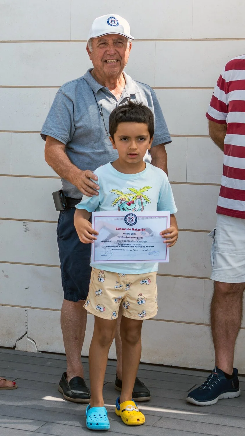 Un niño sostiene un certificado de participación en cursos de natación durante el verano 2023, acompañado por un adulto mayor que lo apoya en un evento al aire libre.