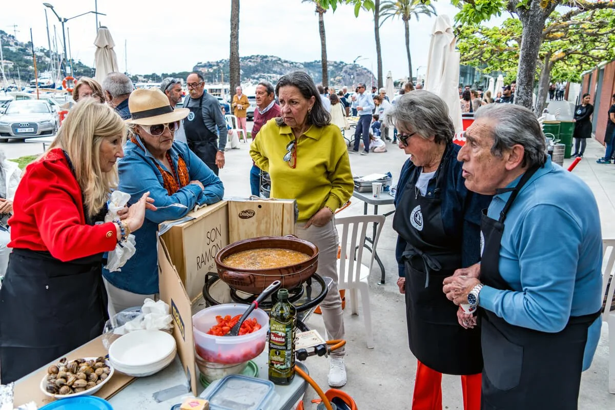 Personas en una reunión en un restaurante o boathouse junto al puerto, probando o vendiendo comida tradicional con una olla grande de sopa o guiso en el centro de la mesa.