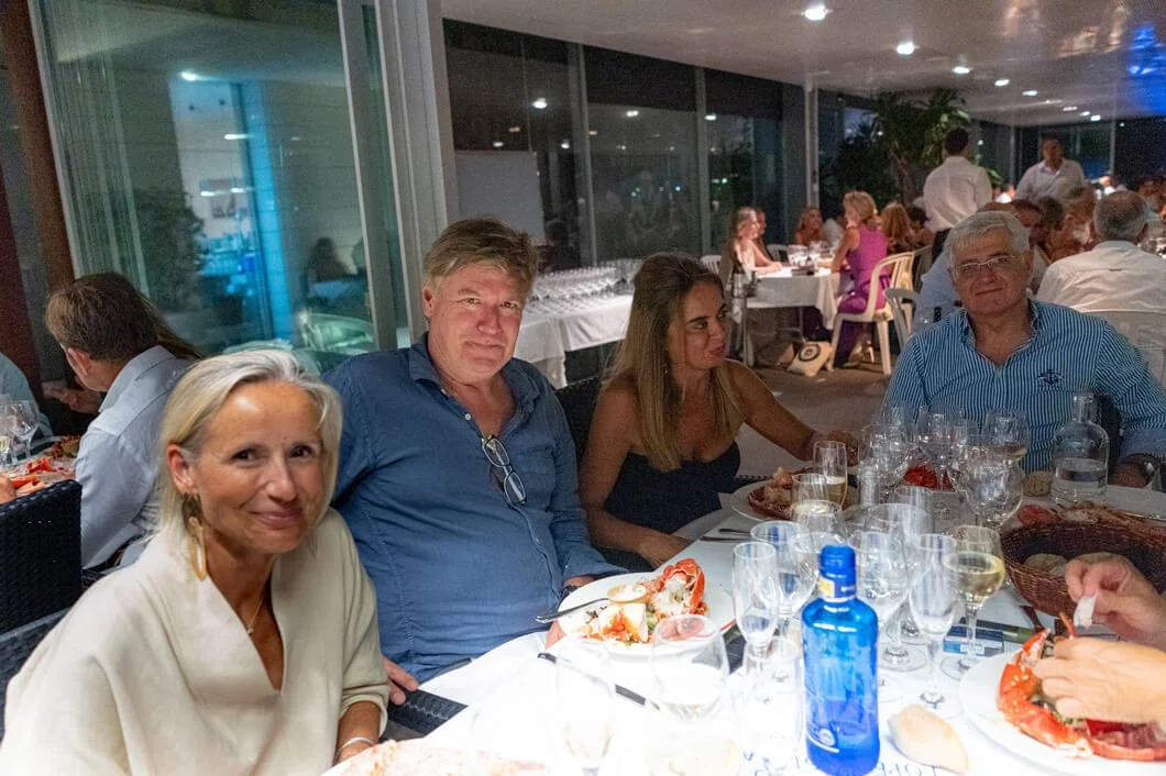 Grupo de personas en una cena en un restaurante, sentados en una mesa con comida y bebida, ambiente elegante y con muchas personas en el fondo.