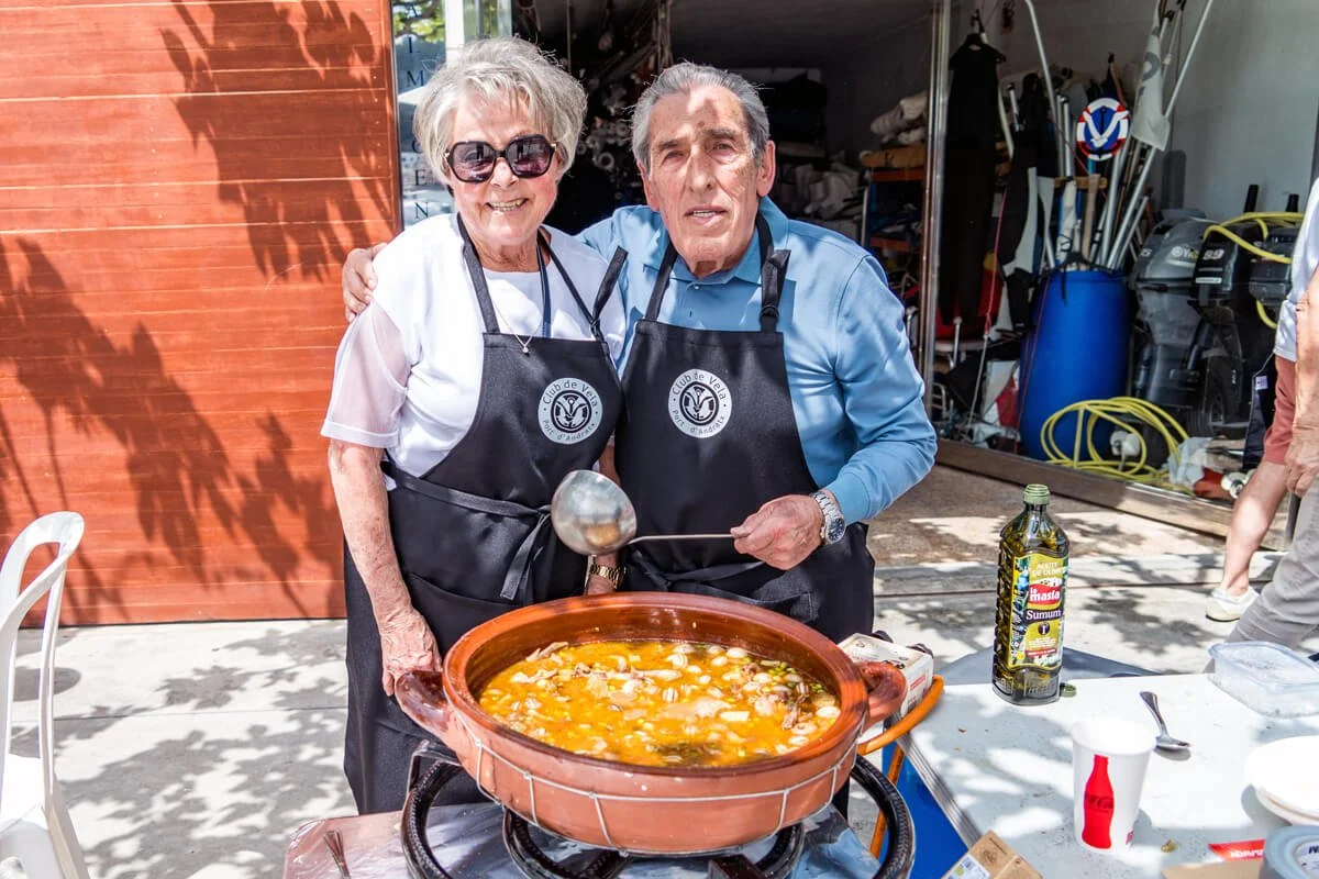 Una pareja mayor con delantal de chef en un evento al aire libre, preparando una sopa en una olla grande de barro.