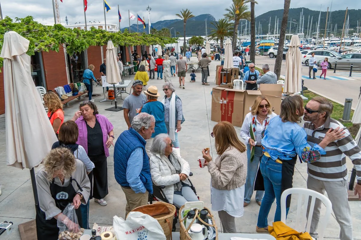 Personas socializando en una plaza cerca del mar con barcos y montañas al fondo, en un día soleado.