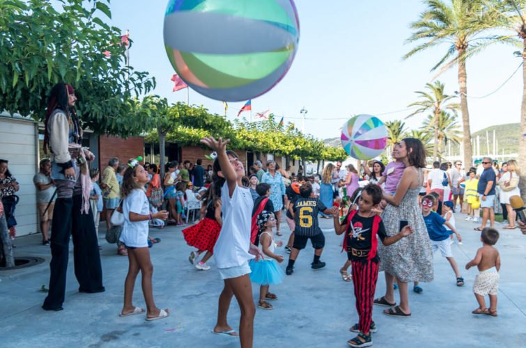 Niños jugando y lanzando un balón grande en un parque con palmeras y muchas personas participando en una celebración