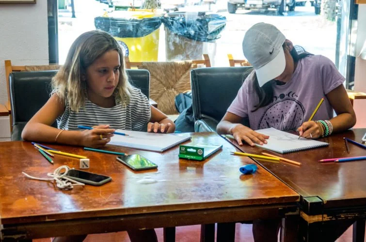 Dos niñas estudiando en una mesa con cuadernos, lápices, y un teléfono móvil en un ambiente interior.