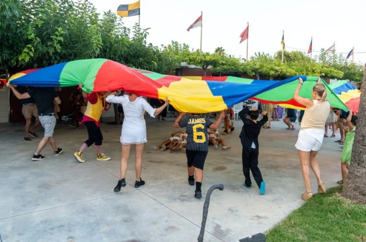 Niños y adultos jugando con un paracaídas de colores en un espacio al aire libre.