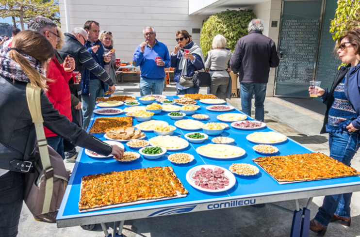 Varias personas sirven y disfrutan de una comida al aire libre frente a una mesa larga con diferentes tipos de pizza y acompañamientos en placas y bowls. 