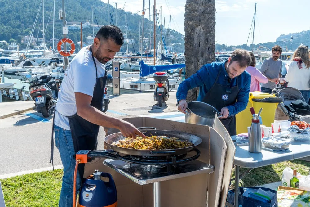 Hombres cocinando comida en un puesto al aire libre cerca del puerto con yates en el fondo.