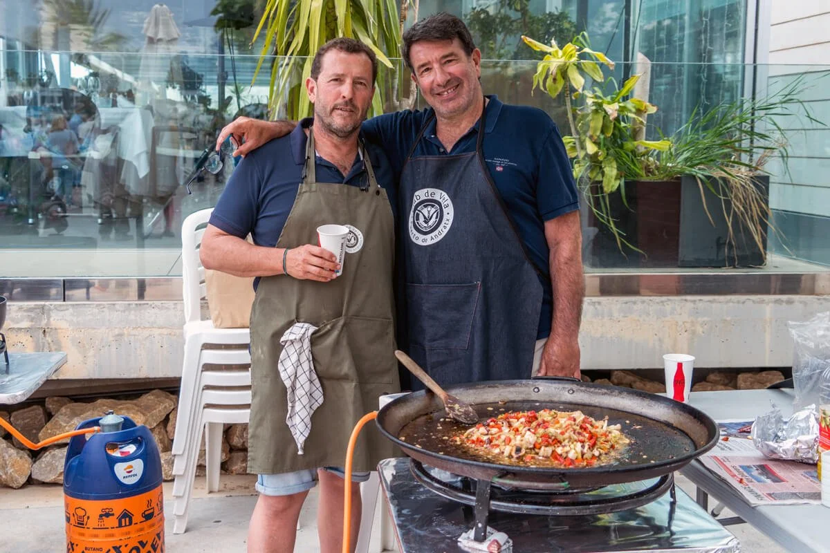 Dos hombres con delantales, uno de ellos sosteniendo una taza, cocinando una pizza en una parrilla gigante en un espacio abierto, con plantas y personas en el fondo.
