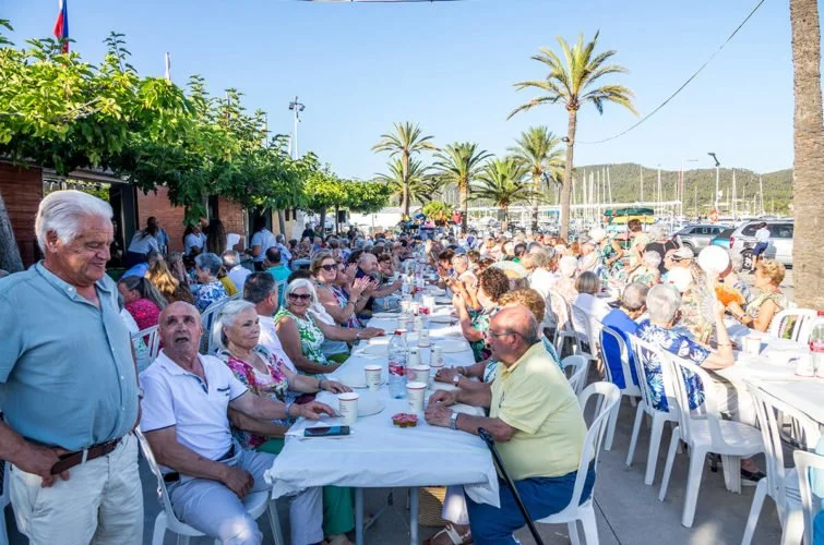 Personas mayores celebrando en una comida al aire libre, en un lugar con palmeras y barcos en el fondo.