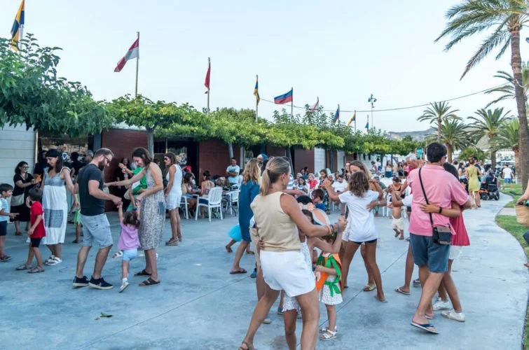 Personas bailando y socializando en un evento al aire libre, con banderas y palmeras en el fondo.