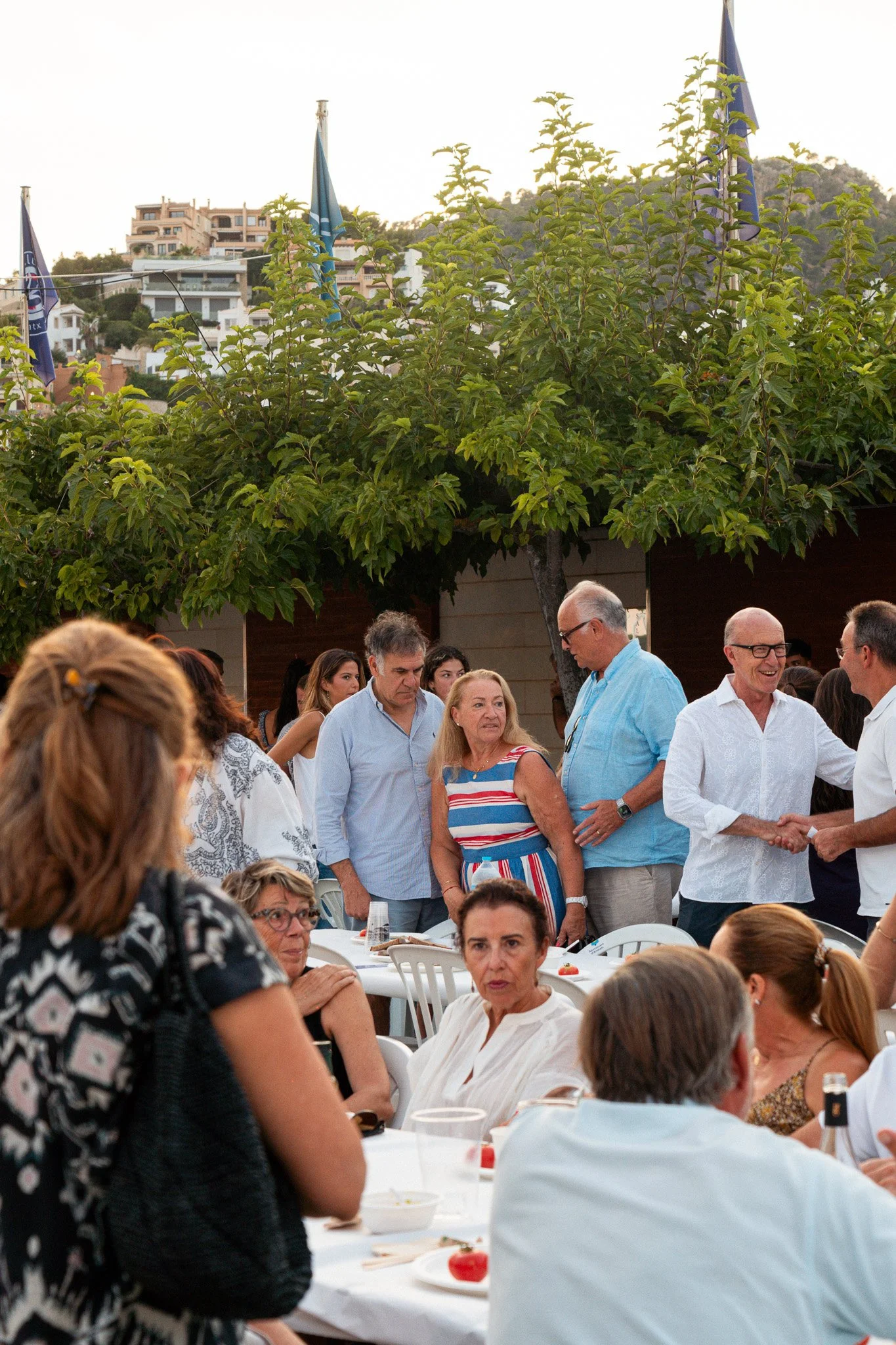Personas reunidas en una comida al aire libre, con mesas y sillas blancas, en un entorno con árboles y edificios en el fondo, durante el atardecer.