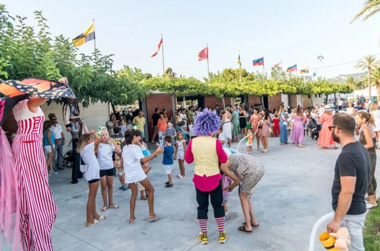 Personajes disfrazados con globos y una multitud de niños y adultos participando en una celebración al aire libre con banderas en el fondo.
