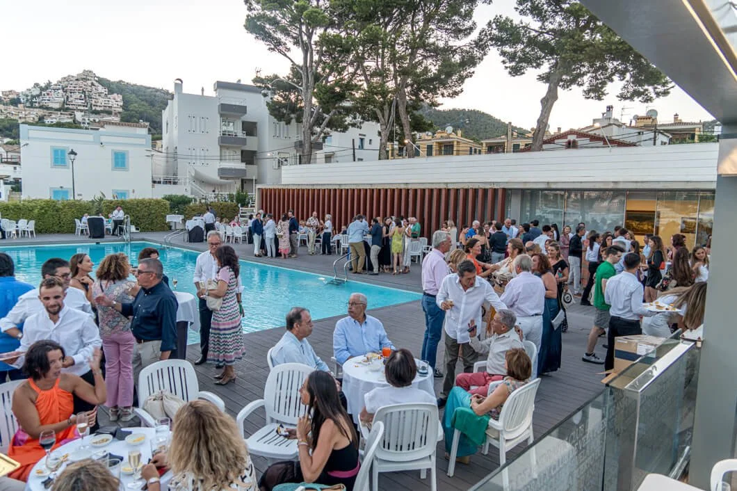 Reunión o fiesta en una piscina con muchas personas socializando, algunas sentadas en mesas y otras de pie, con vista a edificios y montañas al fondo en un ambiente al aire libre.