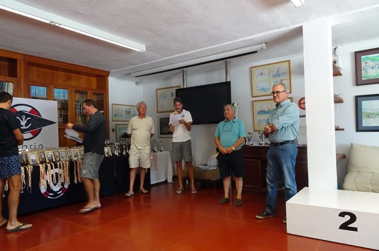 Reunión o premiación en interior con cinco personas, mesa con trofeos y medallas, pantallas y cuadros en las paredes.