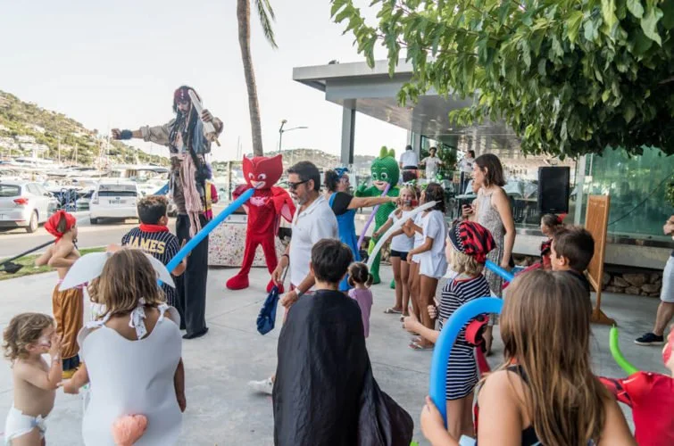 Niños y adultos en una celebración al aire libre con personajes de disfraces de gatos rojo y verde, y globos, en un ambiente festivo y alegre.