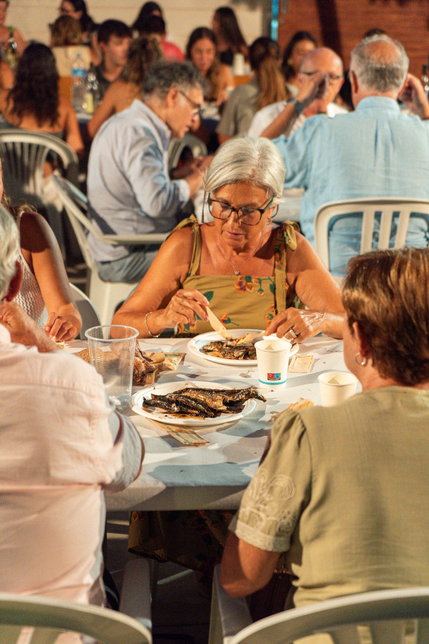 Varias personas sentadas alrededor de una mesa en un evento social, comiendo y conversando, con platos de comida y bebidas sobre la mesa.
