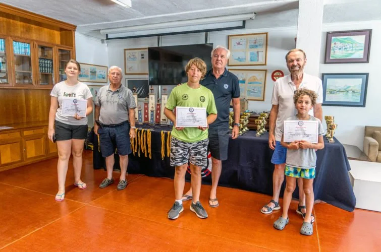 Cinco personas, tres niños y dos adultos, sosteniendo certificados y posando en una sala con mesa de trofeos y adornos, en un acto de premiación.