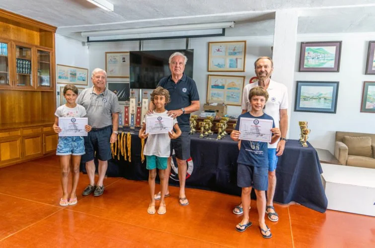 Tres niños sosteniendo diplomas, con tres adultos detrás, en una sala con trofeos y premios en una mesa, decorada con cuadros en las paredes.