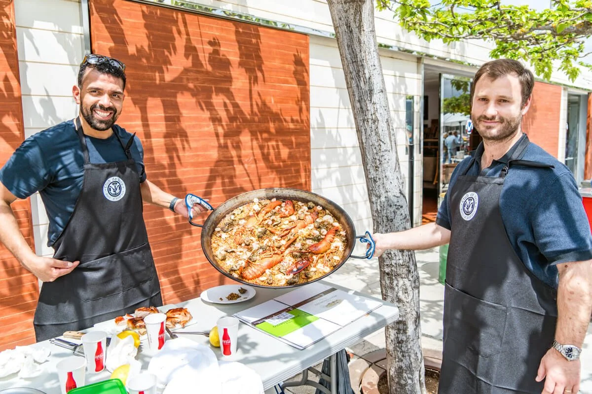 Dos hombres con delantales negros sosteniendo una paella con mariscos en un ambiente al aire libre con árboles y paredes de color terracota.
