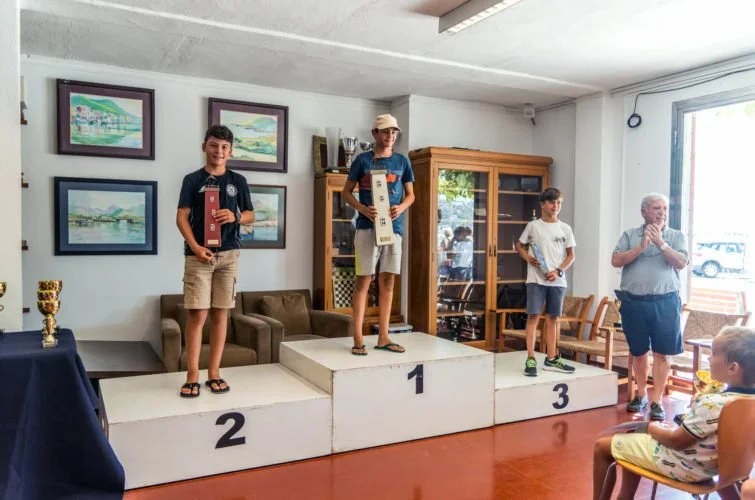 Tres niños en un podio de premios, el primero en el centro, la segunda a la izquierda y la tercera a la derecha, en un entorno interior con un adulto celebrando.