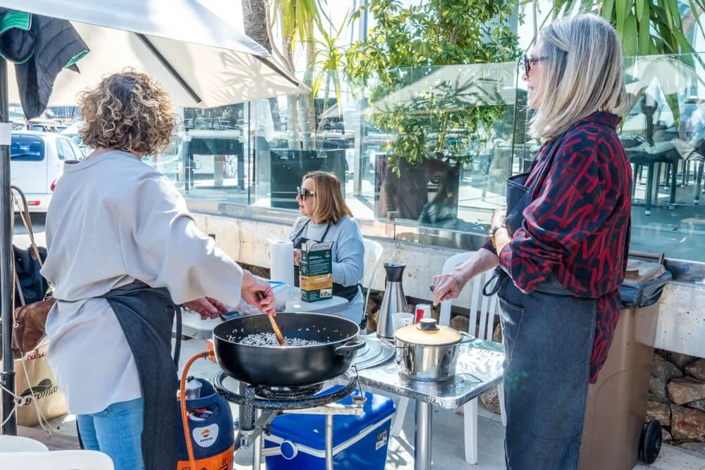 Tres mujeres cocinando y conversando en un espacio abierto con sombra y plantas.