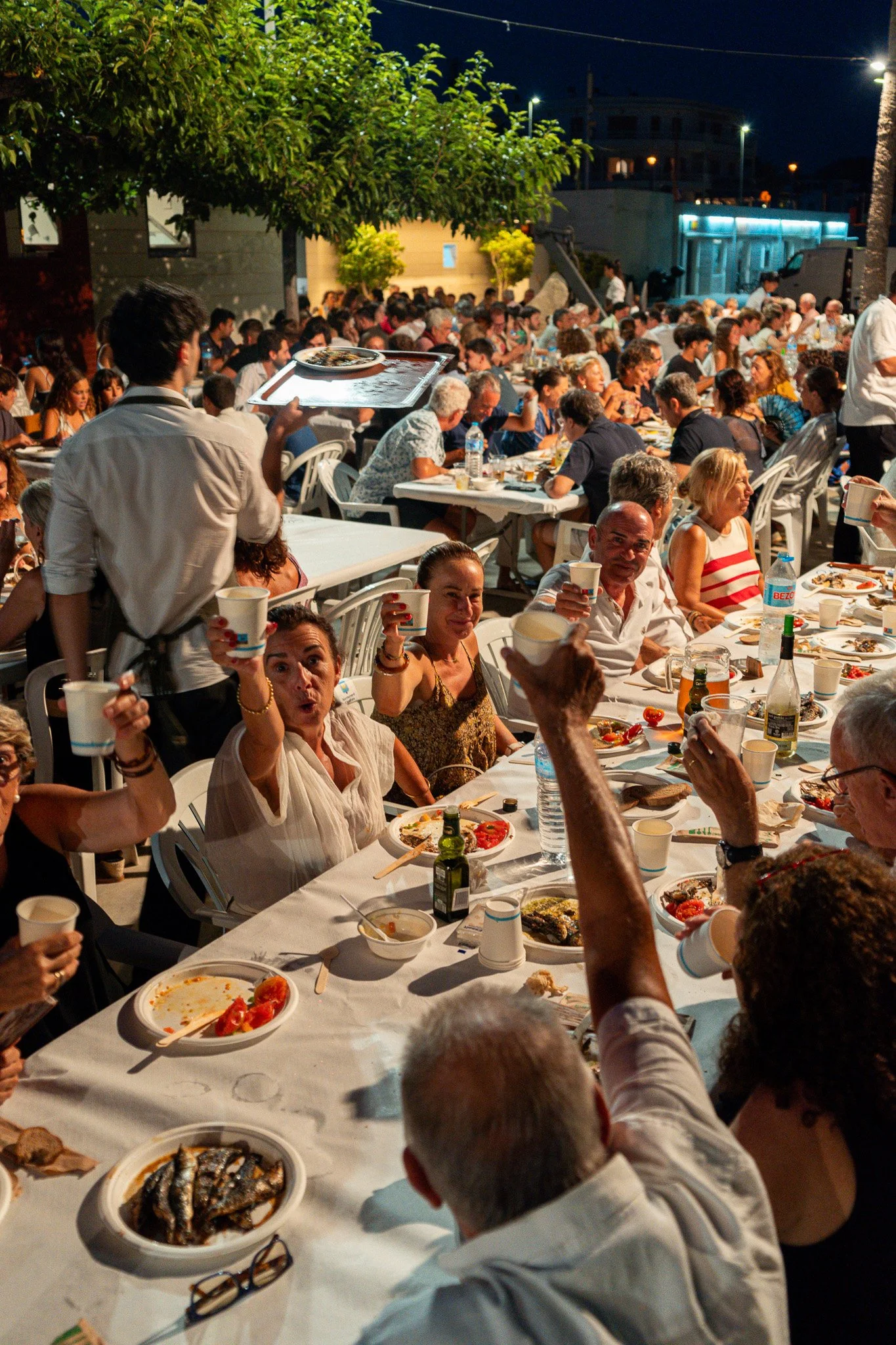 Una escena de una comida o celebración al aire libre por la noche con muchas personas sentadas en mesas, comiendo y disfrutando juntos. El ambiente parece festivo y convivial.