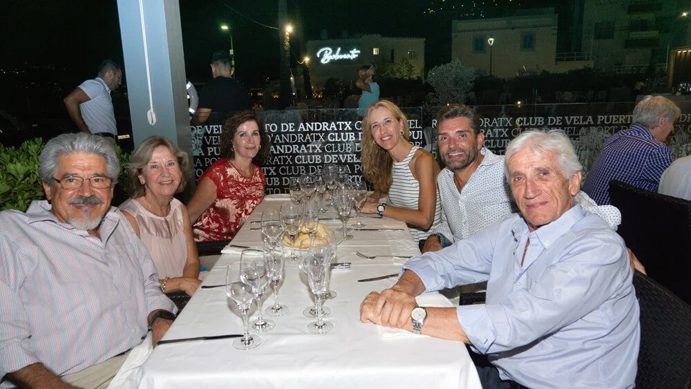 Grupo de personas sentadas en una mesa de restaurante en la noche, algunas sonriendo y con copas de vino y platos con pan, fondo con letras en la pared y luces de la ciudad.