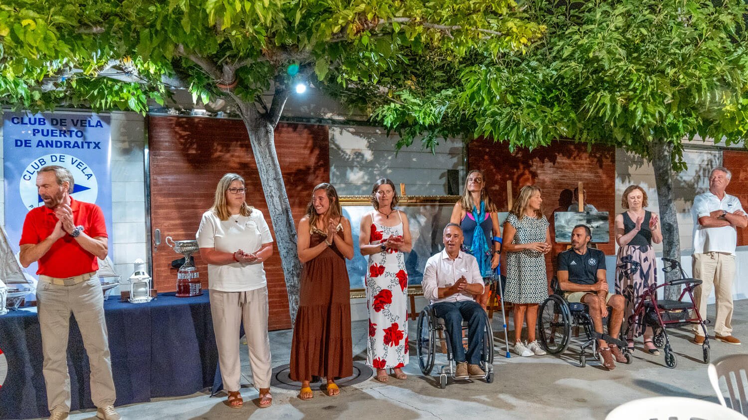 Grupo de personas en un evento al aire libre, algunas en sillas de ruedas, en una noche con árboles y decoraciones, con un banner que dice 'Club de Vela Puerto de Andraix'.