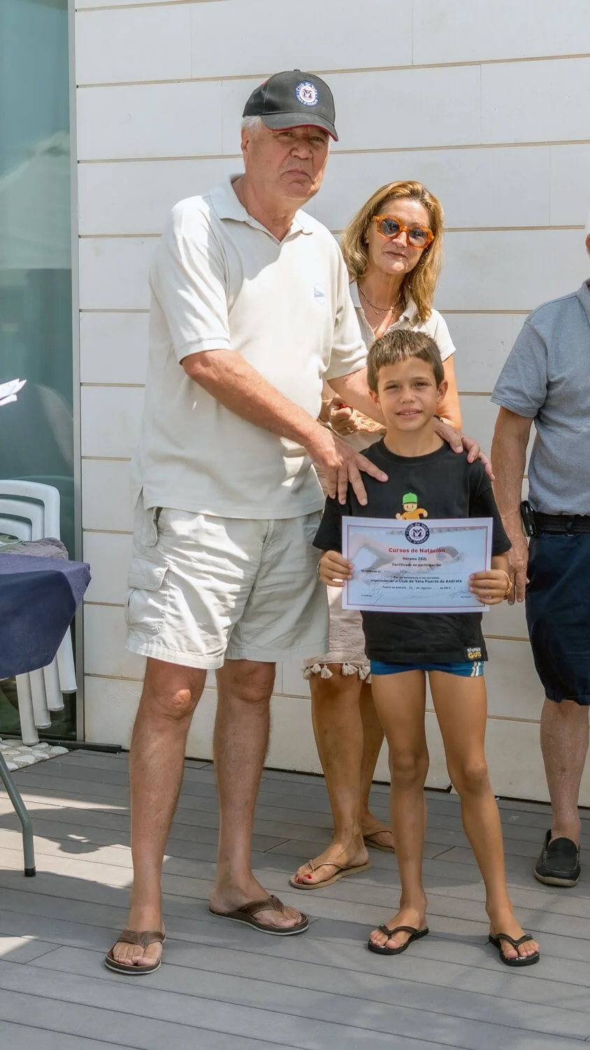 Niño sonriendo sosteniendo un certificado de premiación, acompañado de adultos en una ceremonia al aire libre.