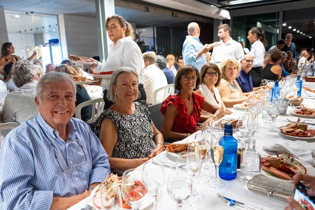 Personas sentadas en una mesa en una cena o comida formal, con platos de comida, copas de vino y agua, en un lugar iluminado y concurrido. Algunas personas están sirviendo y atendiendo a los invitados.