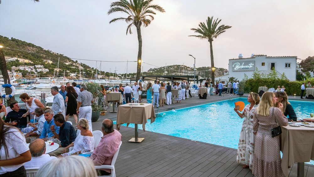 Evento social junto a la piscina con vistas a un puerto y yates, personas conversando y comiendo al atardecer, palmeras y un edificio con letrero de restaurante.
