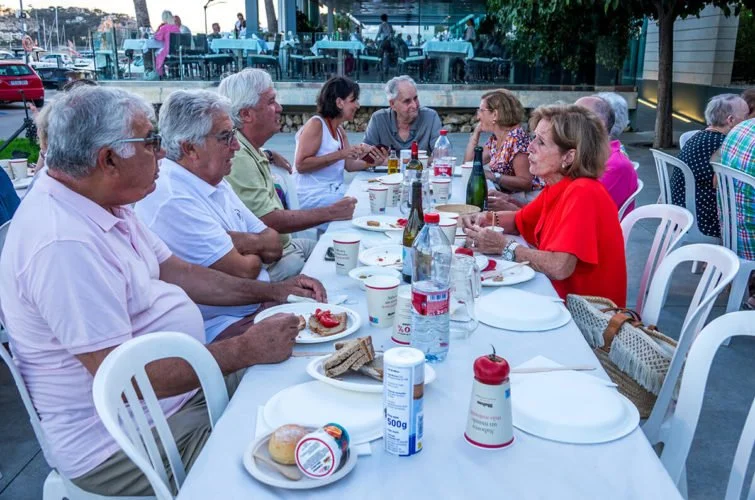 Grupo de personas mayores sentadas en una mesa al aire libre en un restaurante, compartiendo comida y bebidas.
