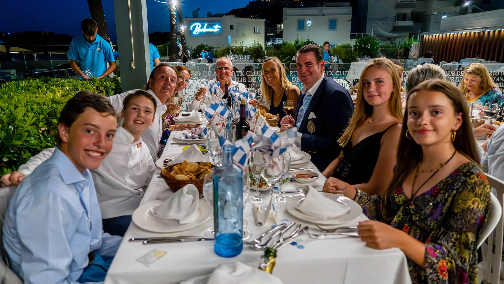 Grupo de personas sentadas en una mesa en una cena al aire libre por la noche, sonriendo y disfrutando de la comida y la bebida, con decoraciones festivas y ambiente alegre.
