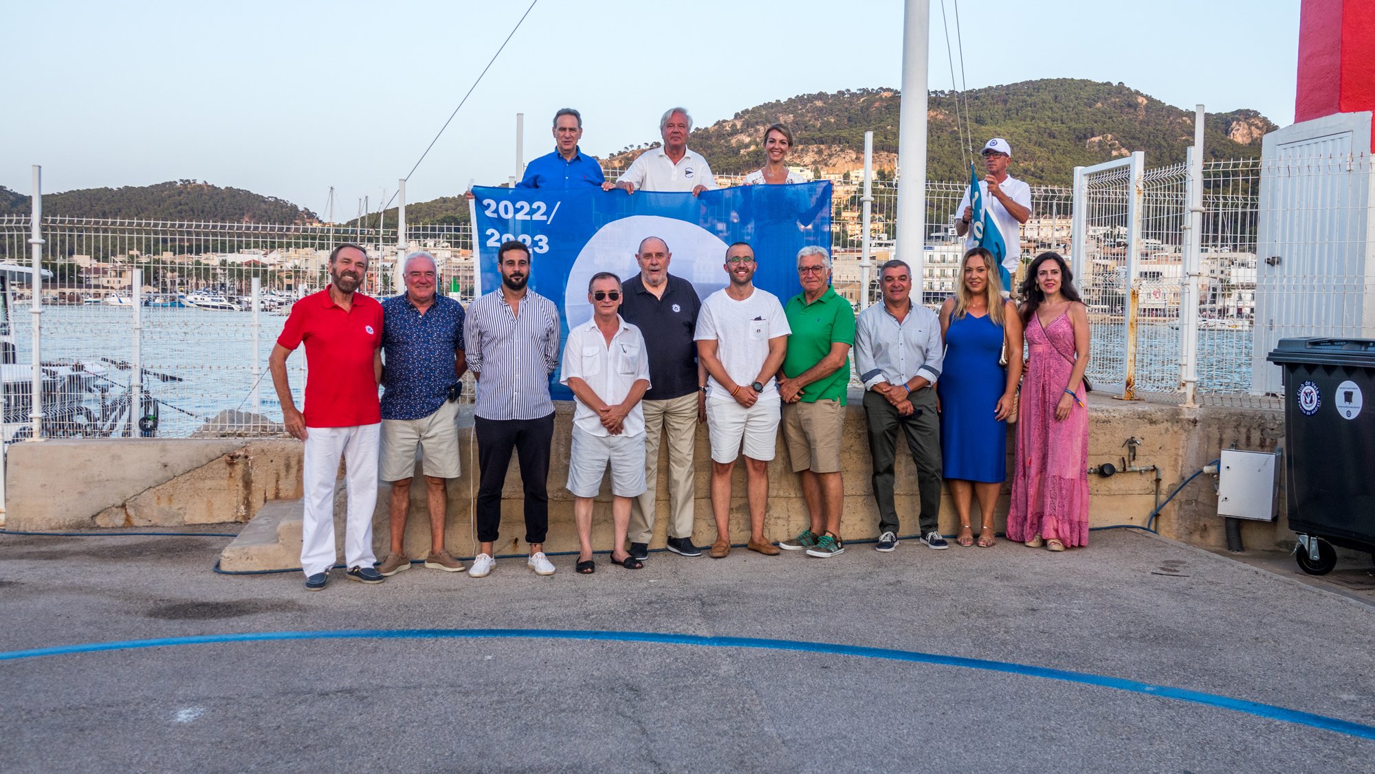 Grupo de varias personas posando frente al mar con un cartel de los años 2022 y 2023, algunos sosteniendo banderas, en un entorno de puerto o marina, con montañas de fondo.